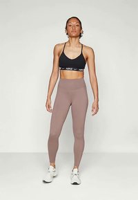 Fekete sportmelltartó fehér Nike logóval, amelyet magas derekú, egyszínű barna leggingsszel párosítanak. A modellen fehér sportcipők vannak.