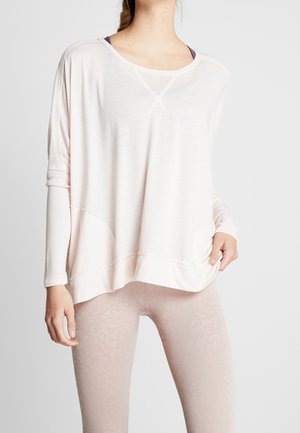 Osoba ubrana w luźną, bladoróżową bluzkę z długim rękawem oraz jasnoróżowe prążkowane legginsy na jednolitym tle.