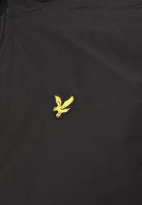Zwarte stof met een gladde textuur, voorzien van een klein, geborduurd geel arendlogo aan de linkerkant. Minimalistisch ontwerp.