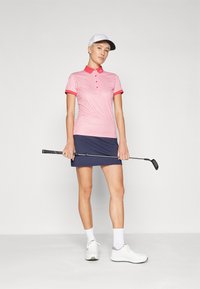 Červeno-biely vzorovaný polo tričko skombinované s námorníckom modrom sukni. Model drží golfovú palicu, má na sebe biele topánky a bielu čiapku.