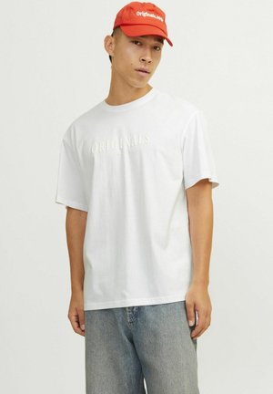 Hvid bomulds t-shirt med "ORIGINALS" tekst i creme. Sammen med lyseblå denim-bukser og en rød cap med hvid tekst.