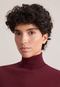 Maglione a collo alto bordeaux a costine con un design aderente. Tessuto morbido e collo alto. Tono della pelle levigato e capelli ricci sono visibili.