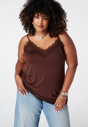 Vrouw met krullend haar die een bruin hemdje met kant en lichtblauwe spijkerbroek draagt, geaccentueerd met ringen, armbanden en een zilveren ketting.