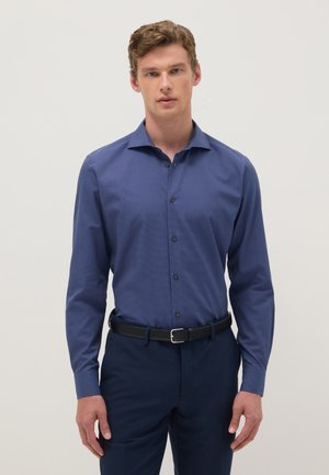 Jeune homme debout, portant une chemise bleue habillée rentrée, un pantalon foncé et une ceinture noire, sur un fond clair uni.