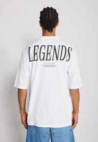 T-shirt blanc à manches courtes avec un grand texte noir "LEGENDS" au dos, accompagné d'un texte plus petit en dessous. Tissu en coton doux, coupe décontractée.