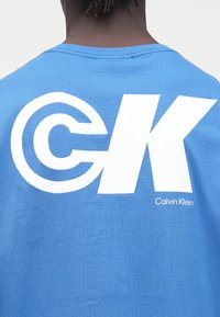 Άτομο που φοράει μπλε πουκάμισο με μεγάλο λευκό λογότυπο "CK" και μικρό "Calvin Klein" τυπωμένο στην πλάτη, πάνω σε απλό φόντο.