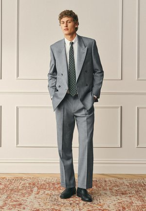Young man standing indoors wearing a gray double-breasted suit, white shirt, patterned black tie, and black shoes on a patterned rug.