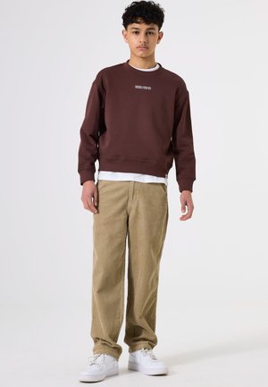 Sudadera marrón con un logo blanco impreso en el pecho, combinada con pantalones de pana beige y zapatillas blancas. Conjunto simple y casual.