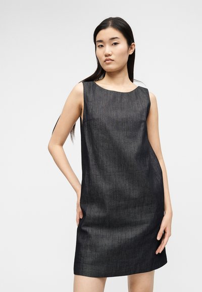 Νεαρή γυναίκα με μακριά σκούρα μαλλιά, που φοράει ένα αμάνικο, σκουρογκρι φόρεμα μέχρι το γόνατο, ποζάροντας μπροστά σε απλό φωτεινό φόντο.