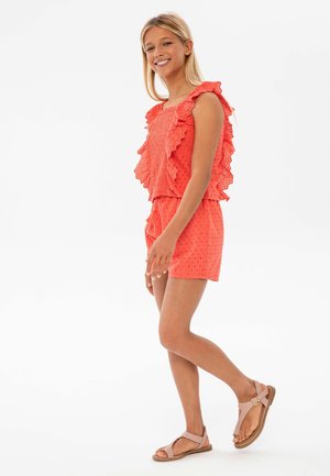 Conjunto sin mangas en color coral con detalles con volantes, textura de ojales y shorts a juego; combinado con sandalias planas de color marrón claro.