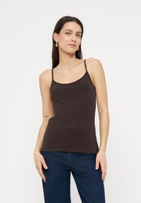 Kvinne med langt mørkt hår som bærer gullfargede hoops, en tettsittende mørk brun spaghetti-stropp topp og mørkeblå jeans, stående med hendene avslappet.