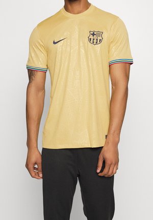Person wearing a light yellow FC Barcelona soccer jersey with multicolor striped sleeve cuffs and black pants, standing against a plain background.