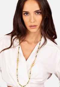 Collana di perline con accenti verdi, gialli e dorati, con perline di forme e dimensioni diverse, indossata sopra una camicia bianca.