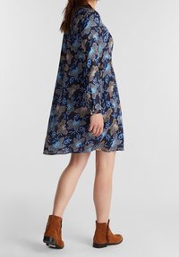 Abito floreale blu navy con maniche lunghe, caratterizzato da motivi multicolori di fiori e foglie. Indossato con stivaletti marroni alla caviglia.