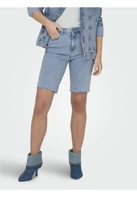 Shorts en denim avec un ourlet brut, lavage bleu clair, associés à des bottines en denim retroussées. Le modèle adopte un look décontracté avec une veste en denim boutonnée.