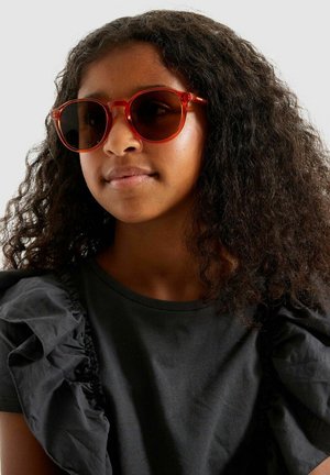 Lunettes de soleil surdimensionnées teintées de rouge avec des verres foncés et une forme ronde, portées avec un t-shirt noir avec des détails froncés aux épaules.