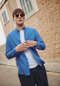 Lichtblauw button-down shirt met lange mouwen, gelaagd over een wit T-shirt. Donkere broek. Zonnebril met ronde monturen. Natuurlijke stenen achtergrond.