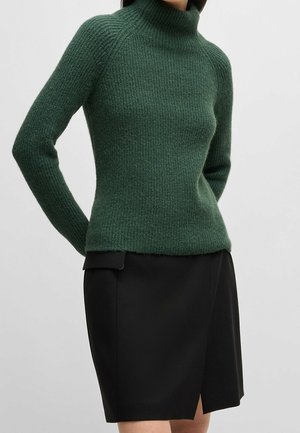 Femme portant un pull à col roulé côtelé vert et une jupe noire longueur genou avec une fente à l'avant, debout devant un fond uni.