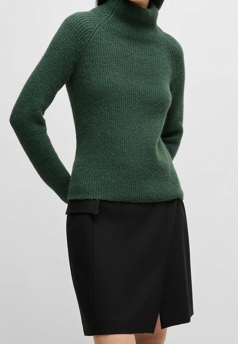 Femme portant un pull à col roulé côtelé vert et une jupe noire longueur genou avec une fente à l'avant, debout devant un fond uni.