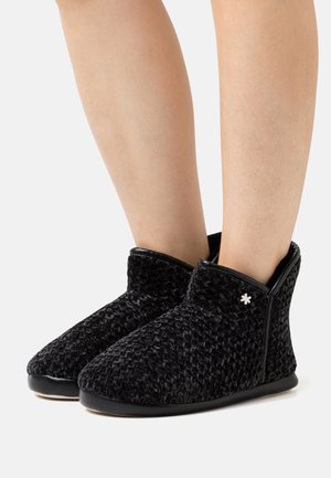 Schwarze, strukturierte Stiefeletten aus weichem Stoff mit einem gewebten Muster, versehen mit einem kontrastierenden, glänzenden Besatz und einem kleinen weißen Akzent.