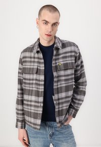 Grå rutete flanellskjorte med knapper, to brystlommer, og en gul logo accenter, iført over en mørkeblå skjorte og kombinert med jeans.