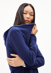 Navyfarbener Kapuzenpullover aus strukturiertem Strick, mit taillierten Ärmeln und umgeschlagenem Kragen. Das Modell hält den Stoff nah am Hals.