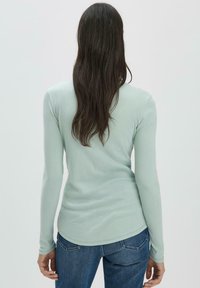 Maglietta a maniche lunghe verde chiaro con orlo arrotondato, texture liscia e design aderente, abbinata a jeans di denim blu scuro.