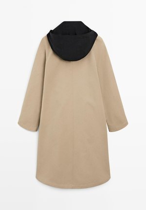 Beige lang frakke med brede ærmer og en sort hætte, vist bagfra på hvid baggrund.