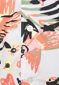 Gros plan sur un tissu blanc imprimé de motifs floraux abstraits en corail, jaune, vert et noir, avec deux boutons blancs le long d'une couture verticale.