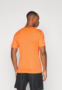 Kurzarmliges, athletisches Shirt in leuchtendem Orange, aus glattem Stoff mit Rundhalsausschnitt und figurbetontem Design, getragen mit schwarzen Shorts.