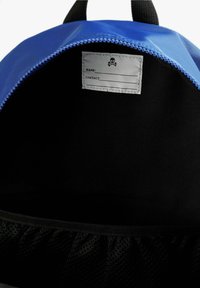 Interior de una mochila azul con un forro negro, que cuenta con una etiqueta para el nombre y la información de contacto, y un bolsillo de malla en la parte inferior.