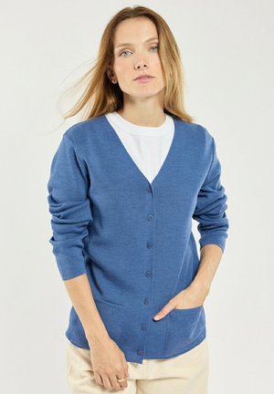 Femme aux cheveux châtain clair portant un cardigan bleu boutonné sur une chemise blanche et un pantalon beige, debout avec une main dans la poche.