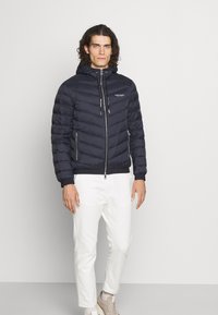 Marineblaue Steppjacke mit Kapuze, gequiltetem Design, vorne mit Reißverschluss und zwei seitlichen Taschen. Kombiniert mit weißen Hosen und beigen Sneakers.