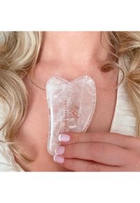 Transparant kristallen gua sha gereedschap in de vorm van een hart, met een glad oppervlak en een gegraveerd logo in roségoud, vastgehouden tussen vingers met roze nagels.