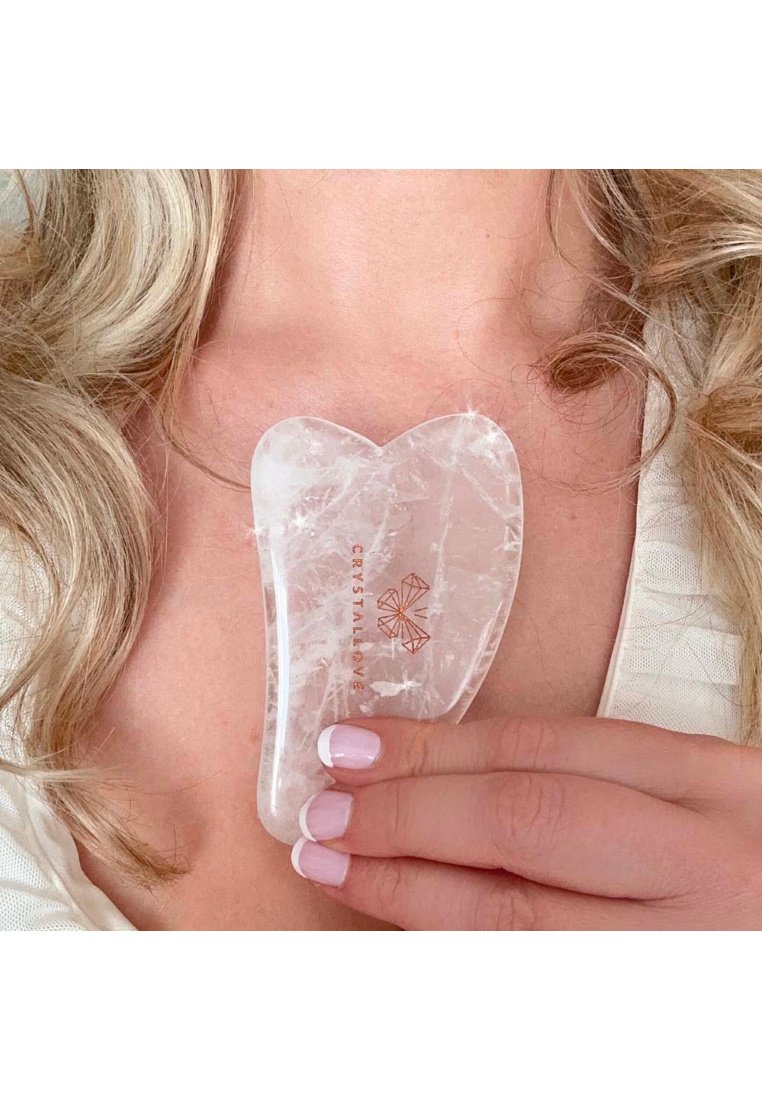 Transparant kristallen gua sha gereedschap in de vorm van een hart, met een glad oppervlak en een gegraveerd logo in roségoud, vastgehouden tussen vingers met roze nagels.