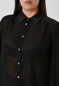 Camicia nera trasparente con colletto, dotata di sei bottoni d'argento lucidi lungo la parte anteriore e piccole pieghe sulle spalle.