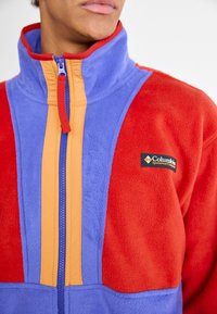Chaqueta de forro polar con paneles multicolores en rojo, azul y naranja, con cuello alto y cremallera frontal. Logo de Columbia en el pecho.