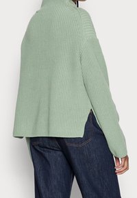 Lichtgroene gebreide trui met een hoge kraag, ribbels, zijsplitten en een losse pasvorm, gecombineerd met donkere denim broek.