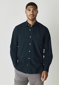 Camicia blu navy con taschino sul petto, maniche lunghe e bottoni bianchi. Il tessuto ha una consistenza liscia. Abbinata a pantaloni grigi chiari.