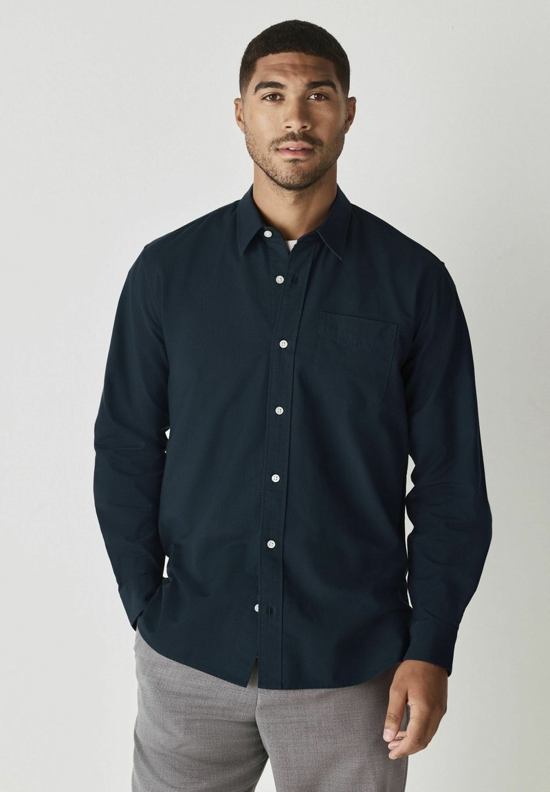 Camicia blu navy con taschino sul petto, maniche lunghe e bottoni bianchi. Il tessuto ha una consistenza liscia. Abbinata a pantaloni grigi chiari.