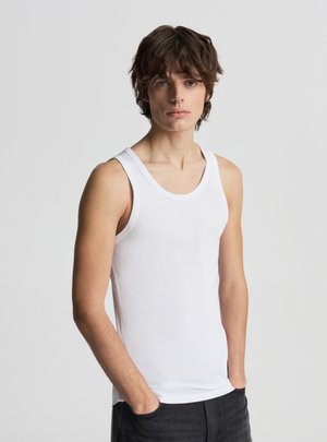 Giovane uomo con capelli castani mossi, indossa una canotta bianca senza maniche e pantaloni neri, in piedi con le mani in tasca davanti a uno sfondo semplice.