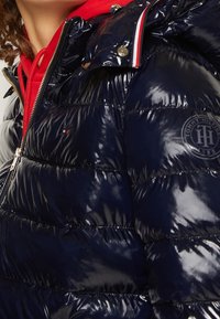 Częściowy widok osoby w błyszczącej granatowej puchowej kurtce z czerwonymi, białymi i niebieskimi detalami oraz czerwoną bluzą z kapturem pod spodem.