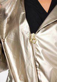 Metalen gouden zip-up jack met een gladde, glanzende textuur. Bevat een hartvormige ritssluiting en een opgevouwen kraag. Zwarte binnenlaag zichtbaar.