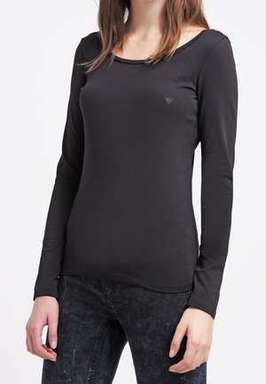 Vrouw die een aangesloten zwart shirt met lange mouwen en donker gewassen jeans draagt, staand tegen een effen witte achtergrond.