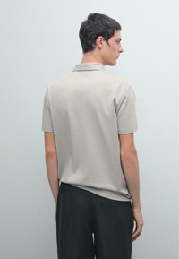 Polo beige con una texture morbida, maniche corte e colletto classico, abbinato a pantaloni scuri, visto da dietro su uno sfondo semplice.