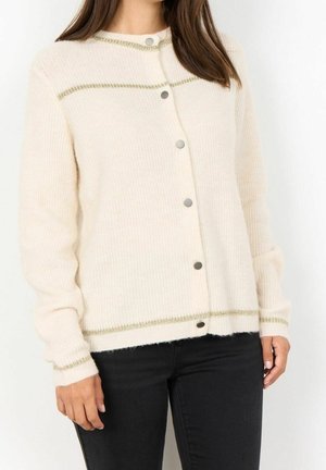 Femme portant un cardigan crème boutonné avec un bord doré et un pantalon noir, debout devant un fond blanc uni.