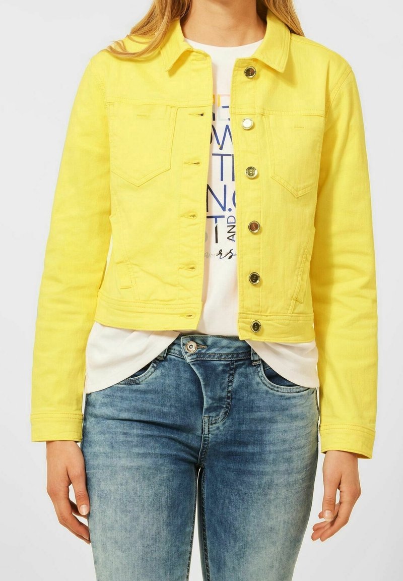 Gelbe Jeansjacke mit Kragen, Fronttaschen und Messingknöpfen, kombiniert mit einem weißen Hemd und hellblauen Jeans. Strukturierter Stoff.