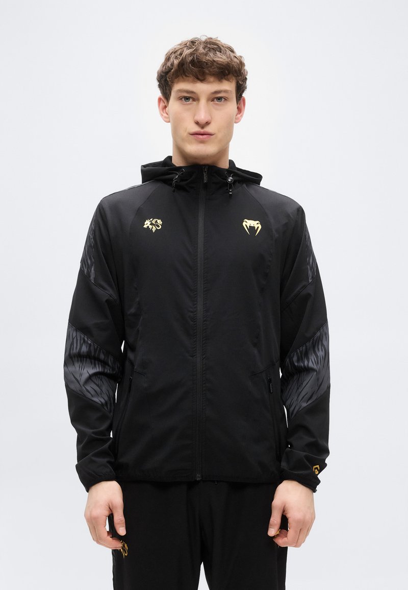 Μαύρο μπουφάν με κουκούλα, χρυσά λογότυπα στο στήθος, υφασμένα μαύρα μανίκια και διπλές πλευρικές τσέπες. Λείο ύφασμα με κομψό σχέδιο.