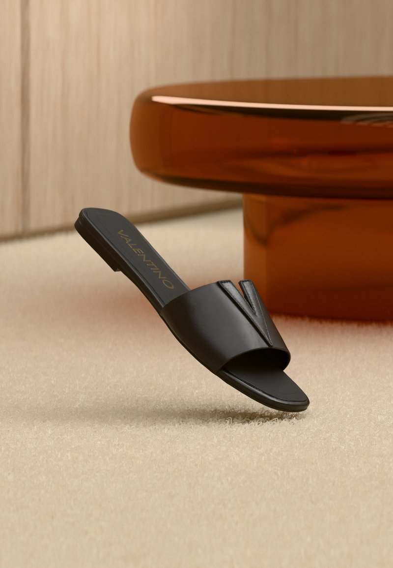 Sandale noire Valentino à bout ouvert posée sur un tapis beige avec une table ronde marron dans une pièce éclairée doucement et neutre.