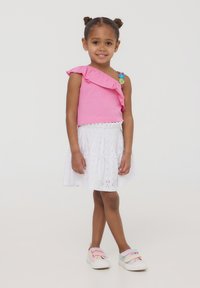 Top rosa a spalla unica con volant abbinato a una gonna bianca in pizzo. Piedi adornati da scarpe slip-on in colori pastello.